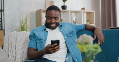 Güzel görünüşlü mutlu, gülümseyen sakallı Afrikalı Amerikalı evdeki yumuşak kanepede oturup arkadaşlarıyla akıllı telefondan sohbet ediyor.