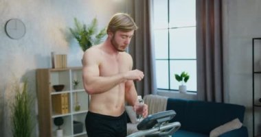 İyi görünümlü, kaslı, aktif sakallı, koşu bandında yürürken evde egzersiz yapan, şişeden su içen ve vücut suyunu yeniden düzenleyen adam.