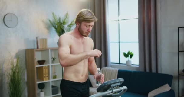 Good-looking fit muscular active bearded guy with naked torso walking on treadmill while exercising at home and drinking water from bottle to replanish body water balance — Stock Video