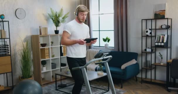 Goed uitziende sportieve actieve bebaarde man in sportkleding joggen op loopband thuis en het houden van video chat met behulp van tablet-apparaat — Stockvideo