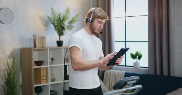 집에서 러닝 머신을 연습하는 동안 헤드셋을 통해 정보를 찾고 있는 건강해 보이는 스포츠 성 수염을 한 남자 — 비디오