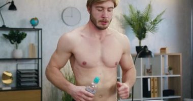 İyi görünümlü, çalışkan, kaslı, sakallı, çıplak gövdeli bir adamın portresi. Koşu bandında koşuyor ve antrenman sırasında şişeden su içiyor.