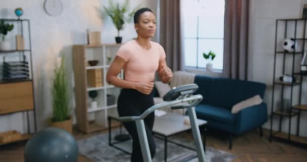 Home ochtend workout concept waar mooie glimlachende fit sportieve Afrikaanse Amerikaanse vrouwelijke persoon in sportkleding kijken in de camera tijdens het lopen op loopband — Stockvideo