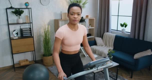 Mooie glimlachende actieve sportieve Afro-Amerikaanse vrouw in training kleding kijken in de camera tijdens het lopen op loopband tijdens de ochtend thuis workout — Stockvideo