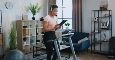 Güzel, kendine güvenen, aktif, kulaklıklı genç bir kadın sabah antrenmanı sırasında evde koşu bandında yürürken tablet üzerinde çalışıyor.