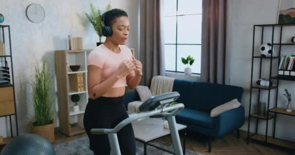 Приємна усміхнена спортивна молода афроамериканка у тренувальному одязі біжить на біговій доріжці і одночасно слухає улюблену музику в навушниках вдома. — стокове відео