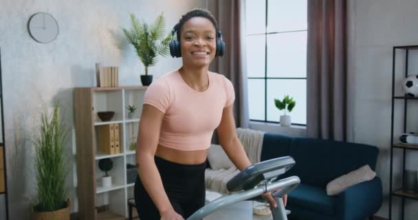 Очаровательная улыбающаяся спортивная активная молодая женщина в тренировочной одежде, бегущая по беговой дорожке, наслаждающаяся музыкой в наушниках и показывающая большие пальцы на камеру — стоковое видео