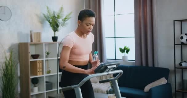 Schöne müde, sportlich schlanke junge Frau in Sportbekleidung trinkt Wasser während des Trainings auf der Laufbahn beim Heimtraining — Stockvideo