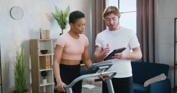 在家里的跑步机上锻炼时，有魅力的年轻女子在留胡子男子手中的平板电脑屏幕上观看 — 图库视频影像