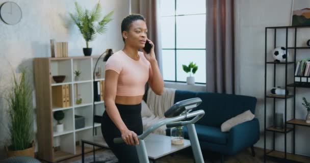 Beautiful smiling active sporty african american woman in training clothes walking on treadmill during home morning workout and talking on phone with friend — Stock Video