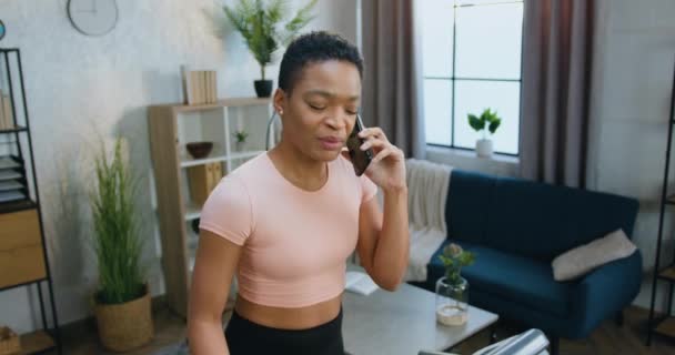 Attrayant souriant actif athlétique femme afro-américaine en vêtements de sport a conversation téléphonique agréable pendant la séance d'entraînement à domicile du matin sur la machine à courir — Video