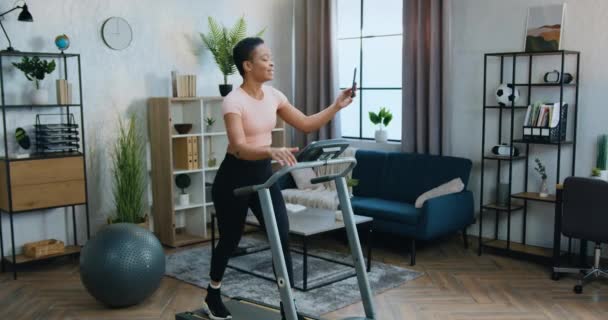 활발히 살아가는 개념, 집에서 육상 경기를 연습하고 휴대 전화로 비디오 채팅을 즐기는, 매력적 인 스포츠적 인 젊은 아프리카 계 미국인 여성 — 비디오