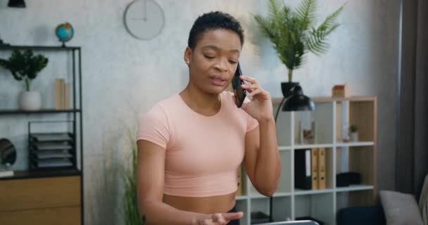 Портрет привлекательной позитивной спортивной подтянутой африканской женщины в светло-розовом топе, который разговаривает по телефону во время упражнений на беговой дорожке дома — стоковое видео