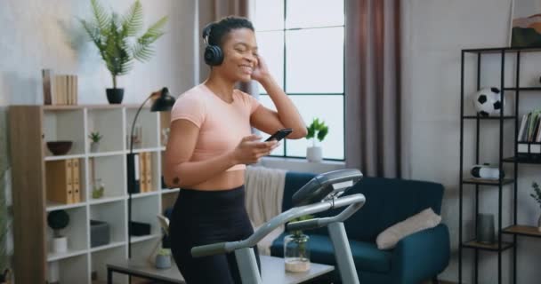 Belle souriante sportive mince femme afro-américaine en vêtements de sport profiter de la musique préférée dans les écouteurs pendant l'entraînement à la maison sur la machine à courir — Video