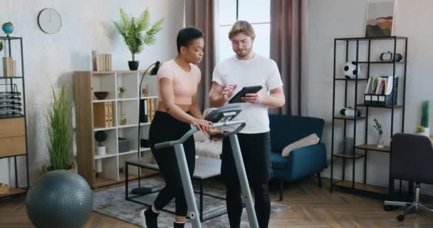 Likable sporty active diverse couple training together on treadmill using special fitness instruction on tablet device at home,healthy lifestyle concept — Stock Video