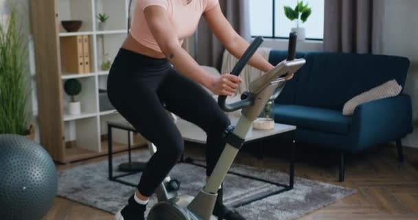 Привлекательный усталый спортивный стройная молодая африканская женщина в тренировочной одежде поворачивая педали на велосипеде упражнения во время домашней тренировки, концепция активного образа жизни — стоковое видео