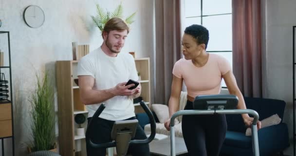 Conceito de treino em casa onde atraente sorrindo casal multiétnico esportivo exercitando juntos na esteira e bicicleta de exercício e falando sobre fotos conjuntas em caras móveis — Vídeo de Stock