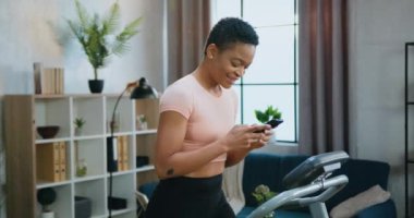 Ev ödevi konsepti. Çekici, neşeli, narin, Afro-Amerikan kadın spor kıyafetleri içinde koşu bandında yürürken telefonda arkadaşlarından haber okuyor.