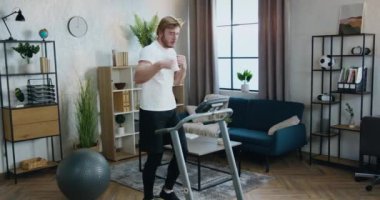 Spor giyim kuşamında sportif sakallı, ev antrenmanı sırasında koşu makinesinde yürüyüş egzersizleri yapan iyi bir sporcu.
