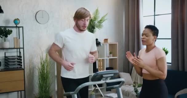 Good-looking active sporty muscular bearded guy in sportswear running on treadmill while his fit african american female friend supporting him with applauses — Stock Video