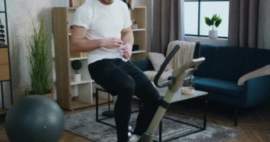 Kapalı mekanda bisiklet konsepti. Evde staj yaparken giysi giyip su içen yakışıklı, aktif, kaslı, sakallı bir adam.