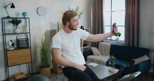 Atrakcyjny uśmiechnięty, aktywny, brodaty facet w odzieży sportowej ćwiczący na rowerze podczas porannego treningu w domu i robiący selfie na swoim smartfonie — Wideo stockowe