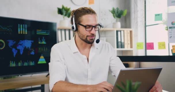 Millennial caucásico masculino sentarse en el lugar de trabajo en auriculares mirando a la computadora portátil durante hablar con el cliente mediante chat web remoto, aplicación de videoconferencia en línea. — Vídeos de Stock