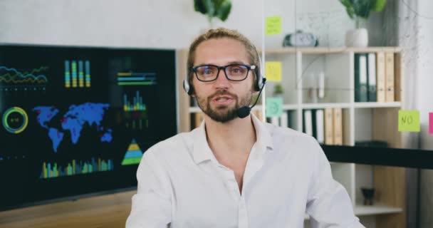 자신감 있는 남자의 얼굴 사진, 책상에 앉아 있는 흰색 셔츠를 입고 헤드폰을 쓰고 사무실에서 웹 카메라로 통화하는 서비스 담당자. — 비디오
