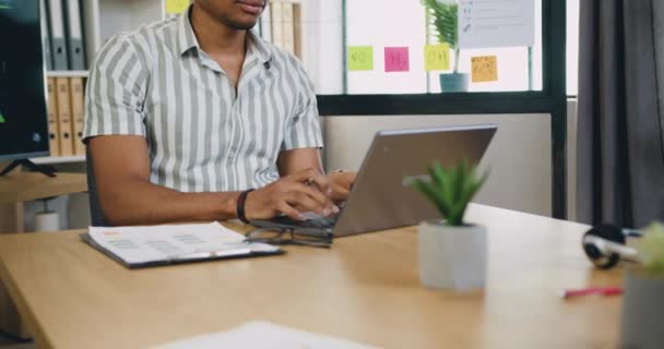 Gericht jonge Afrikaanse man zit in kantoor kamer en met behulp van laptop computer, notities zitten op het werk bureau. — Stockvideo