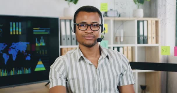 Nahaufnahme Porträt eines afrikanisch-amerikanischen Mannes mit Brille im Hemd, Anrufer tragen drahtloses Headset und schauen in die Kamera im modernen Büroraum — Stockvideo