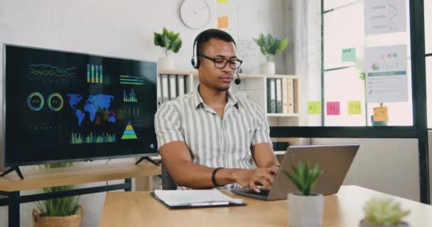 Впевнений молодий африканський бізнесмен носить навушники, слухаючи навчальний курс на конференції масштабування, змушує нотатки сидіти за робочим столом в сучасній офісній кімнаті — стокове відео