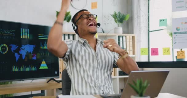 Retrato de joven afro-americano alegre empleado en auriculares sentado en su escritorio y el uso de la computadora portátil en la habitación de la oficina — Vídeos de Stock