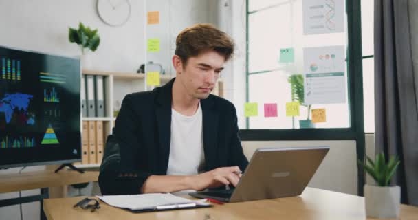 Συγκεντρωμένος νεαρός επιχειρηματίας που εξετάζει την πληκτρολόγηση οθόνης laptop σερφάροντας στο διαδίκτυο ή δουλεύοντας εξ αποστάσεως στο σύγχρονο γραφείο — Αρχείο Βίντεο
