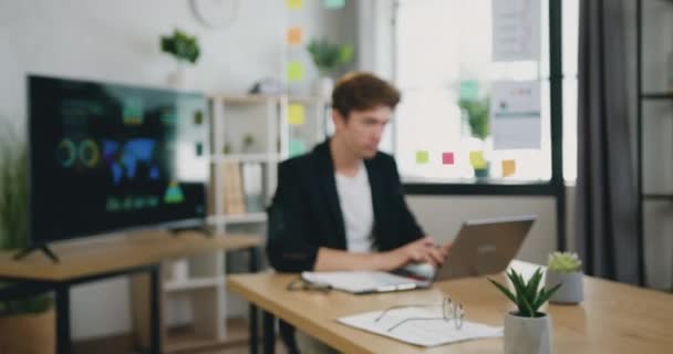 Portret skupionego młodego biznesmena pracującego z laptopem i papierowymi dokumentami w nowoczesnym biurze, uśmiechniętego patrzącego w kamerę. — Wideo stockowe