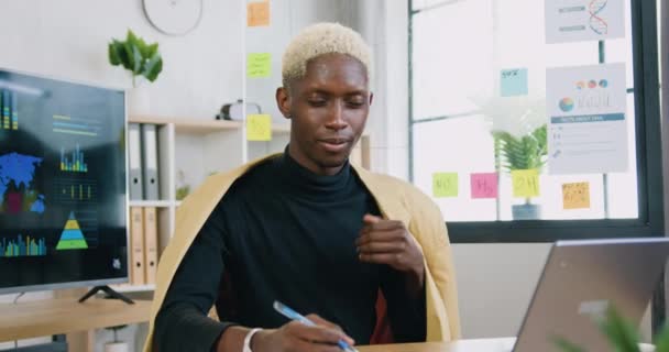 Mavi gözlü, beyaz saçlı, dijital bilgisayarın yanında oturan ve modern oda ofisinde baş parmağını gösteren kameraya gülümseyen Afrikalı yakışıklı adamın portresi. — Stok video