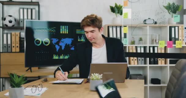Portrét soustředěného mladého podnikatele pracujícího s notebookem a papírovými dokumenty v moderní kanceláři. — Stock video