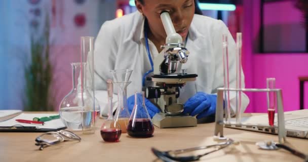 Munkafolyamat az orvosi szobában, ahol nagyon okos, magabiztos afro-amerikai női vegyész kutatja a biológiai mintákat mikroszkóp alatt este — Stock videók