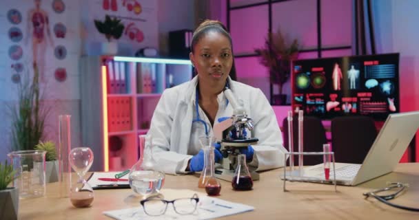 Hermosa confiada joven afroamericana joven químico en uniforme sentado delante de la cámara en el laboratorio de la noche, vista frontal — Vídeo de stock