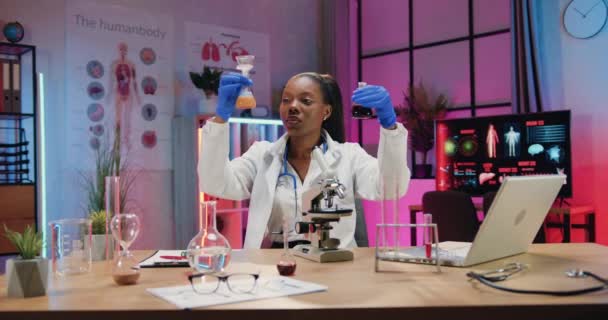 Attraktive selbstbewusste afrikanisch-amerikanische Wissenschaftlerin in Uniform mit medizinischen Handschuhen, die ein Experiemtn mit Flüssigkeiten durchführt und die Ergebnisse in einen Papierbericht schreibt — Stockvideo