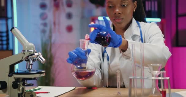 Cute confident high-skilled african american woman scientist in white coat and gloves conducting chemical experiments with liquids and mixing them in flask,front view — Stock Video