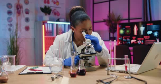 Hermosa mujer química afroamericana trabajadora concentrada que aprende muestras biológicas bajo el microscopio y escribe resultados en un informe de papel en un laboratorio nocturno — Vídeos de Stock