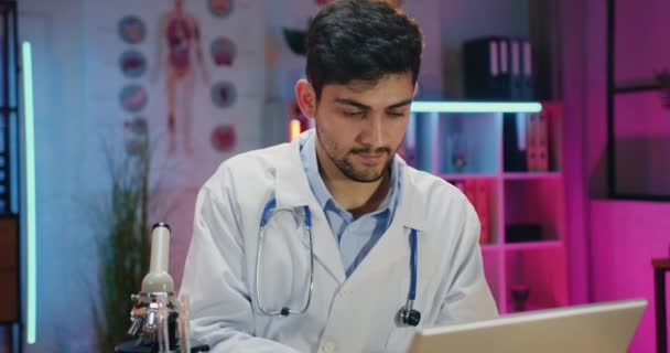 Pravděpodobný kvalifikovaný mladý vousatý vědec v uniformě pracující s mikroskopem a počítačem při práci přesčas ve večerní laboratoři — Stock video