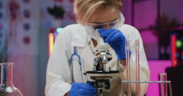 Atractivo concentrado experimentado observante mujer rubia química en máscara protectora, gafas y guantes de aprendizaje de muestras bajo el microscopio en el laboratorio nocturno — Vídeos de Stock