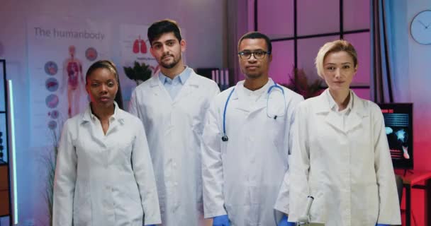 Goed uitziende glimlachende zelfverzekerde hoogopgeleide multi-etnische mannelijke en vrouwelijke artsen in labjassen poseren op camera met gekruiste armen in modern avondlab — Stockvideo