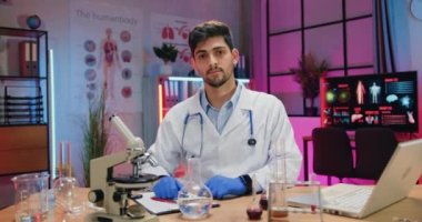 Çekici, kendine güvenen, tıbbi üniformalı ve eldivenli yetenekli kimyager akşam laboratuarında kameranın önünde oturuyor ve iyi bir sembol gösteriyor.