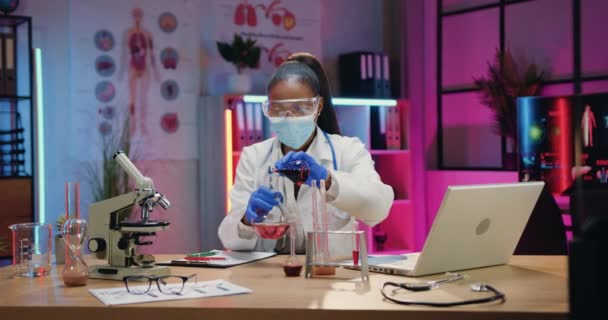 Bella concentrata specializzata donna afro-americana chimico in uniforme e guanti protettivi e maschera facciale condurre esperimento chimico con liquidi in laboratorio sera — Video Stock