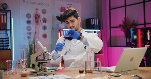 Handsome purposeful qualified bearded chemist in white coat and gloves working with chemical liquids and glass flasks in evening laboratory,front view — Stock Video