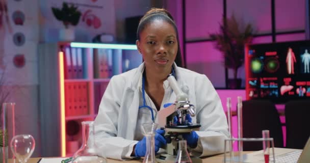 Ziemlich positive erfolgreiche 30-jährige afrikanisch-amerikanische Forscherin im weißen Kittel posiert an ihrem Arbeitsplatz im medizinischen Labor vor der Kamera, Zeitlupe — Stockvideo