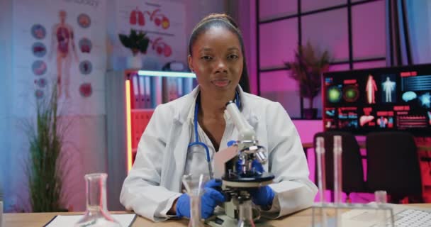 Привлекательный позитивный уверенный умный африканский ученый-американец в медицинской одежде позирует на камеру в вечерней лаборатории и показывает большие пальцы вверх — стоковое видео
