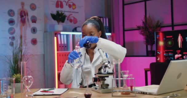 Attractive satisfied responsible skilled african american female chemist in white coat and gloves adding fluid into flask with water and mixing during work in evening lab — Stock Video